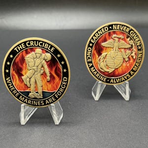 May include: Two gold-coloured challenge coins on clear stands. One coin features a soldier carrying another, with text reading "THE CRUCIBLE WHERE MARINES ARE FORGED." The other coin displays an eagle, globe, and anchor with the text "ONCE A MARINE ALWAYS A MARINE NEVER GIVEN EARNED."