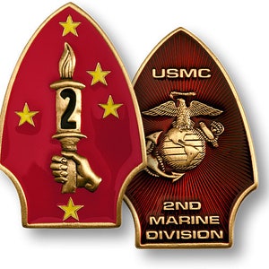 May include: A gold-coloured metal pin with a red and gold enamel design. The pin features a torch with the number "2" on it, surrounded by five gold stars. The bottom of the pin reads "USMC 2ND MARINE DIVISION".