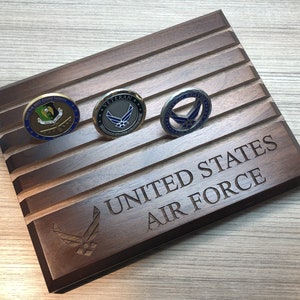 May include: A dark brown wooden display case with three military challenge coins. The case is engraved with "UNITED STATES AIR FORCE" and the Air Force emblem. The coins have various designs and colours.