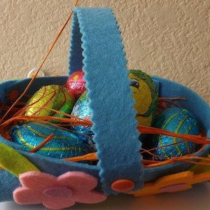 Può includere: Un cestino di Pasqua in feltro blu con manico, riempito di uova di cioccolato avvolte in carta stagnola colorata e dettagli in feltro arancione e giallo.