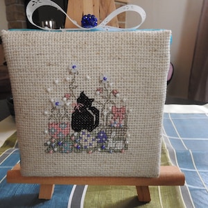 Peut inclure: Une broderie au point de croix représentant un chat noir sur une toile de lin beige avec un ruban bleu et blanc et une perle bleue sur un chevalet en bois. La broderie représente un chat noir assis dans un jardin avec des fleurs et un panier d'œufs.