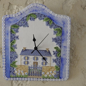May include: A blue and white clock with a cross-stitch design of a house with a fence and flowers. The clock has a white background and black hands.