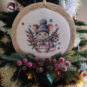 Peut inclure: Ornement de Noël en point de croix avec une souris portant un bonnet et une écharpe bleus, tenant un cadeau. L'ornement est encadré dans un cercle en bois et entouré de verdure festive, de baies rouges et de pommes de pin. Accroché à un sapin de Noël.