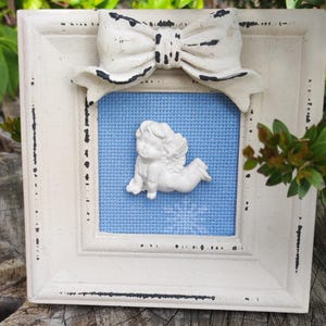 May include: A white frame with a distressed finish and a white bow on top. Inside the frame is a blue background with a white cherub figurine.
