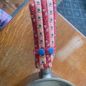 May include: A red, white, and blue floral patterned pot handle cover. The cover is made of fabric and has two blue buttons. The handle cover is attached to a stainless steel pan.