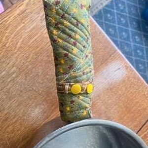 May include: A quilted green and yellow fabric pot handle cover with a floral pattern. The cover is secured with a yellow button closure. The handle cover is resting on a wooden surface next to a metal bowl.