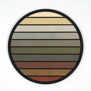 May include: Circular wooden wall art with a black frame. The artwork displays horizontal stripes in a range of earthy colours: beige, grey, olive green, and brown. The design is simple and modern, suitable for home decor.