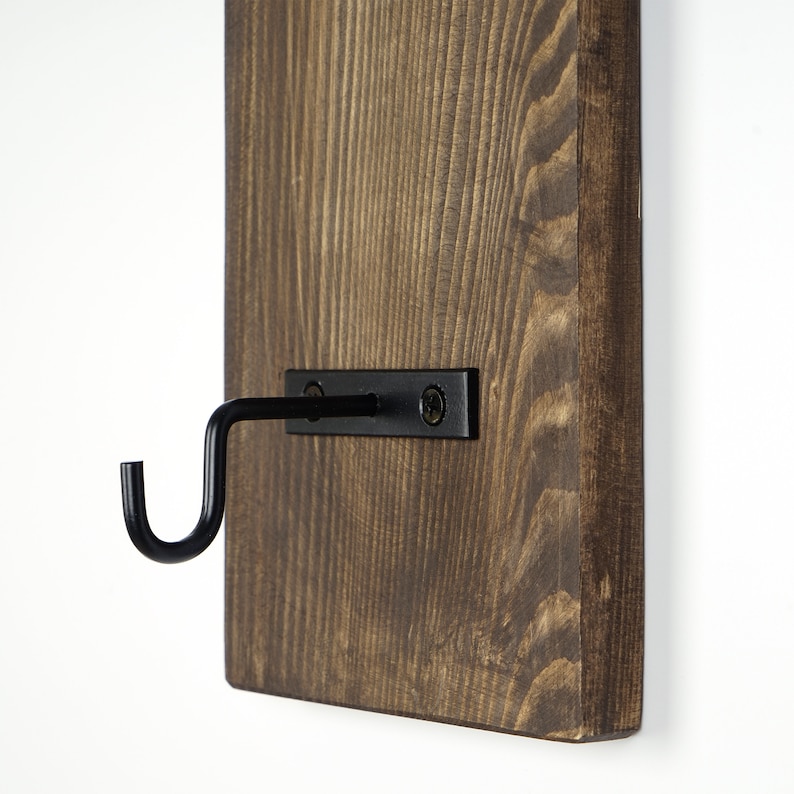 May include: A dark brown wooden wall mounted hook with a black metal hook. The hook is attached to the wall with two black screws.