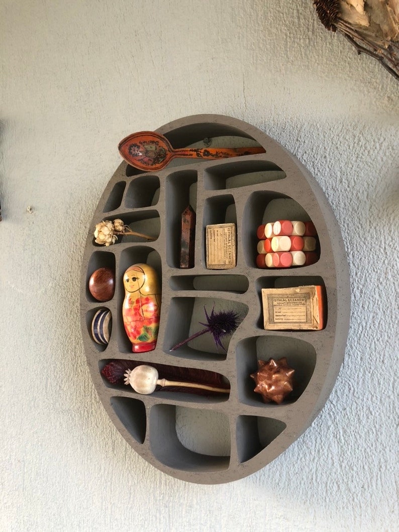 Op de afbeelding: Een ronde, grijze, aan de muur gemonteerde plank met meerdere vakken. De plank is gevuld met verschillende decoratieve items, waaronder een houten lepel, een nestpop, een klein boeket gedroogde bloemen, een kristal, een doosje lucifers en een klein houten beeldje.