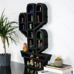 May include: A black wooden cactus-shaped shelf unit with multiple compartments. The shelves display various crystals, small bottles, and a Matryoshka doll. The unit is mounted on a white wall, with a potted plant and books nearby.