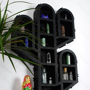 May include: Black wooden cactus-shaped shelf with multiple compartments, displaying various crystals and small glass bottles. The shelf is mounted on a white wall, with a potted plant and a Matryoshka doll in the foreground.