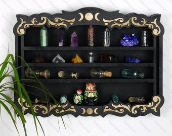 Può includere: Mensola in legno nero con dettagli dorati e decorazioni ornate. La mensola ha tre livelli ed è piena di vari cristalli, piccole bottiglie e un set di bambole russe. La mensola è decorata con un motivo di luna e stelle.
