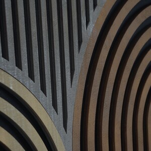 Op de afbeelding: Een close-up van een houten wandkunstwerk met een geometrisch patroon van concentrische cirkels en parallelle lijnen. De cirkels zijn in tinten bruin en beige, terwijl de lijnen zwart zijn.