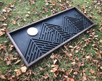 May include: A rectangular wooden wall art piece featuring a mountain design. The artwork has a dark gray background with a geometric mountain silhouette. A light-colored circular element is positioned near the mountains. The frame is a dark brown color.