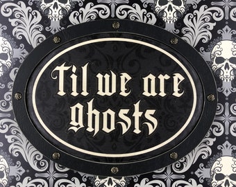 Może przedstawiać: Owalny znak z napisem "Til we are ghosts" gotycką czcionką. Znak jest czarny z białym obramowaniem i otoczony czarnym, skóropodobnym obramowaniem z ozdobnymi ćwiekami. Tło przedstawia powtarzający się wzór czaszek i kwiatowych wzorów.