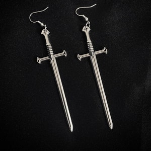 Pode incluir: Um par de brincos prateados em forma de espada. Cada brinco apresenta um design detalhado de espada com uma guarda em forma de cruz e uma lâmina longa e pontiaguda. Os brincos penduram-se em ganchos simples, sobre um fundo escuro.