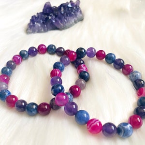 May include: Two beaded bracelets with a variety of colorful stones in shades of blue, purple, and pink. A cluster of amethyst crystals sits in the background. The bracelets are on a white, fluffy surface.