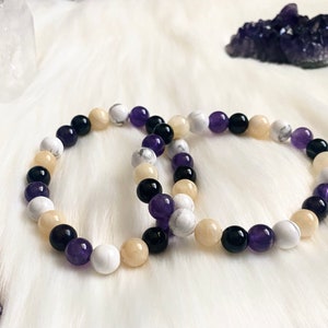 May include: Two beaded bracelets with a mix of colors including white, black, purple, and beige. The bracelets are made of round beads and are displayed on a white, fluffy surface. A cluster of purple crystals is in the background.