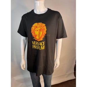 May include: Black t-shirt featuring a gold and orange graphic of a face with the text "VERSACE PROFUMI" in gold. The shirt is displayed on a mannequin. The t-shirt is a crew neck style.