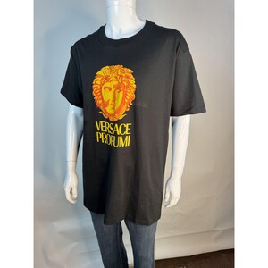 May include: Black t-shirt featuring a gold and orange graphic of a face with the text "VERSACE PROFUMI" in gold. The shirt is displayed on a mannequin. The t-shirt is a classic crew neck style.