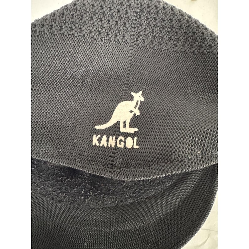 May include: Dark gray Kangol hat with a white kangaroo logo and the brand name. The hat has a textured knit pattern and a curved brim. The logo is centered on the front of the hat.