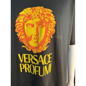 May include: Black t-shirt featuring a yellow and orange Medusa head graphic. The text "VERSACE PROFUMI" is printed in yellow below the graphic. The shirt has short sleeves.