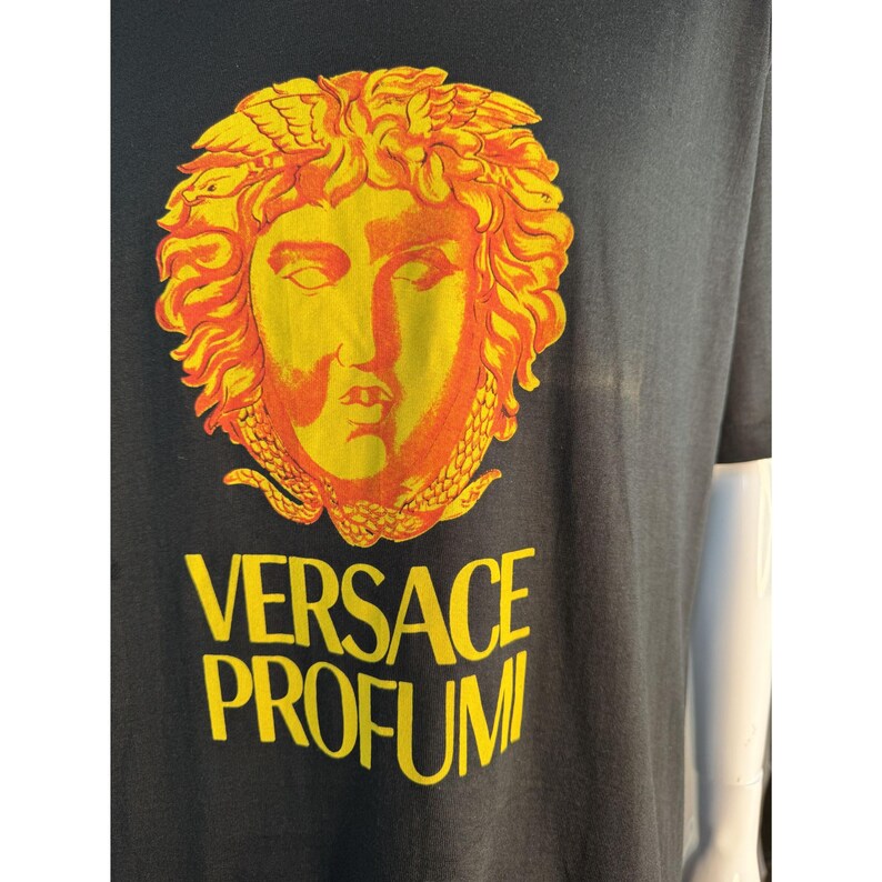 May include: Black t-shirt featuring a yellow and orange Medusa head graphic. The text "VERSACE PROFUMI" is printed in yellow below the graphic. The shirt has short sleeves.