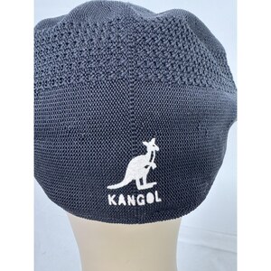 May include: A dark gray Kangol beret hat. The hat features a white kangaroo logo and the brand name "KANGOL" embroidered on the front. The hat has a textured, woven design.