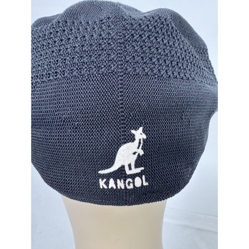 May include: A dark gray Kangol beret hat. The hat features a white kangaroo logo and the brand name "KANGOL" embroidered on the front. The hat has a textured, woven design.