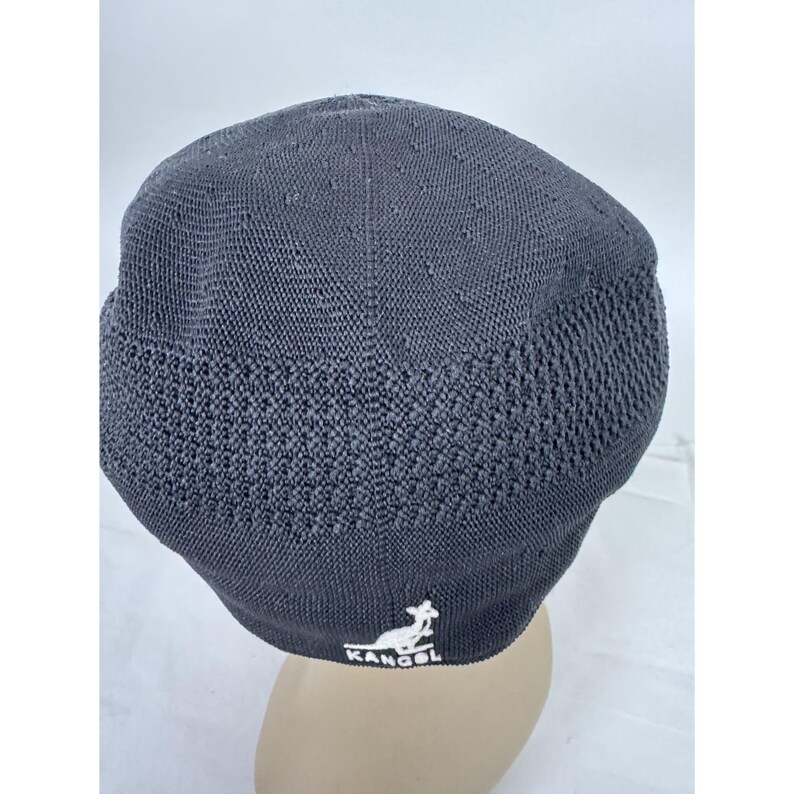 May include: A charcoal gray Kangol Tropic 504 Ventair cap. The cap features a textured knit design with small ventilation holes on the top. The Kangol logo is embroidered in white on the front. This hat is a stylish accessory.