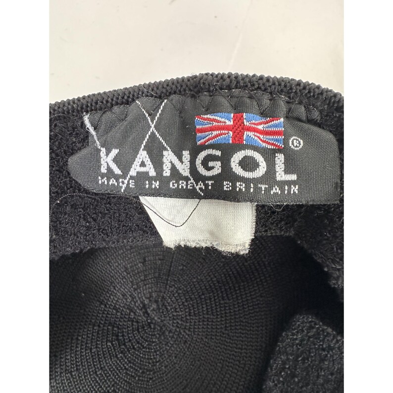 May include: Black Kangol hat with a textured design. The label reads "KANGOL MADE IN GREAT BRITAIN" with the Union Jack flag. The hat is a classic style, suitable for various occasions.