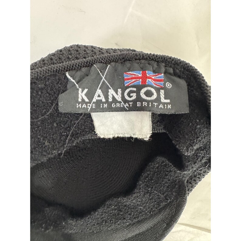 May include: Black Kangol hat with a textured knit design. The label reads "KANGOL MADE IN GREAT BRITAIN" with the Union Jack flag. The hat is a classic style, suitable for various occasions.