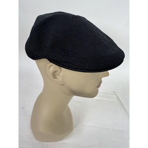 May include: Black flat cap with a textured knit design. The cap is displayed on a mannequin head. The cap has a rounded crown and a short, stiff brim. The cap is a solid black color.