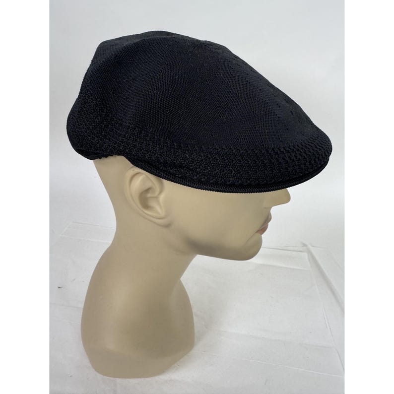 May include: Black flat cap with a textured knit design. The cap is displayed on a mannequin head. The cap has a rounded crown and a short, stiff brim. The cap is a solid black color.