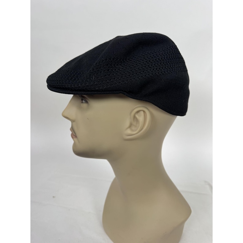 May include: A black flat cap with a textured, breathable design. The cap is made of a woven material and features a short brim. The hat is displayed on a mannequin head against a white background.