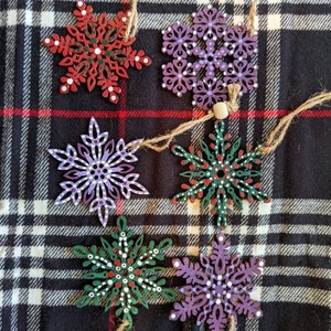 May include: Six wooden snowflake ornaments with painted details. The snowflakes are in various colors, including red, green, purple, and white. They are hanging from twine on a black and white plaid background.