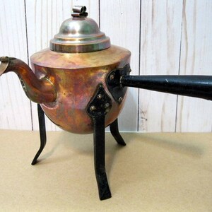 May include: A copper teapot with a black handle and three black metal legs. The teapot has a domed lid with a decorative finial.