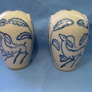 May include: A pair of off-white ceramic salt and pepper shakers. Each shaker features a blue painted design of a deer and foliage. The shakers have a rounded shape with a small opening at the top for dispensing.