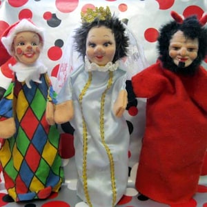 May include: Three vintage cloth dolls: a jester in a colorful diamond pattern, a bride in a white dress with gold trim, and a devil in a red robe with black gloves and horns. The dolls have painted faces.