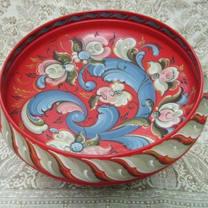 May include: A red wooden bowl with a hand-painted floral design. The bowl features blue, green, white, and pink flowers and swirls. The rim of the bowl is decorated with a gray and red striped pattern.