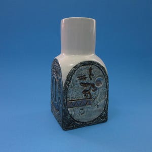 May include: A ceramic vase with a white top and a square base. The vase features a textured design with blue and gray accents, and a decorative oval with what appears to be Asian characters.