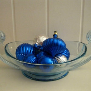 May include: A clear glass bowl with a light blue tint, filled with blue and silver Christmas ornaments. The bowl has decorative handles with a spiral design. The ornaments have ribbed and smooth surfaces.