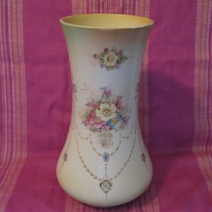 May include: A tall, cream-colored ceramic vase with a floral design. The vase has a wide base and a narrow neck. The floral design features pink, yellow, and blue flowers with green leaves. The vase is decorated with a gold chain pattern.