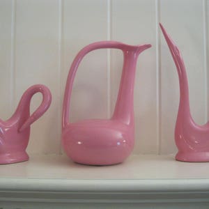 May include: Three pink ceramic swan figurines. Two of the swans have their necks curved into a graceful S shape, while the third swan has its neck extended upwards. The swans are all in a similar style, with a smooth, glossy finish.
