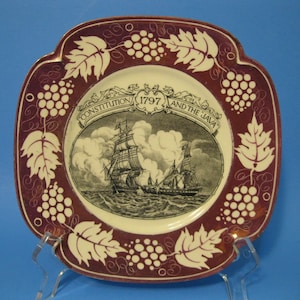 May include: A burgundy ceramic plate with a white rim and a grapevine design. The center of the plate features a black and white illustration of the USS Constitution and the USS Java, with the text "Constitution 1797 and the Java".