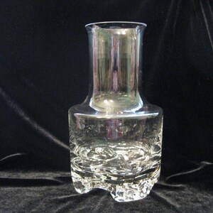May include: A clear glass vase with a wide, textured base and a cylindrical upper section. The base has a unique, sculpted design, and the vase tapers slightly towards the top. The glass reflects light, creating a shimmering effect. The vase is approximately 8 inches tall.