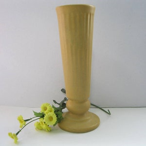 May include: A tall, yellow ceramic vase with a fluted design and a pedestal base. The vase is empty and there are yellow flowers on the table next to it.