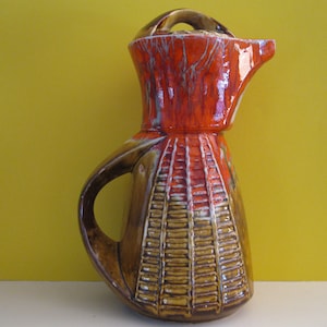 May include: A brown and orange ceramic pitcher with a lid. The pitcher has a textured design and a unique shape.