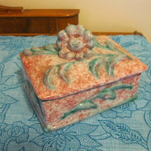 May include: A pink and green ceramic trinket box with a floral design. The box has a raised flower on the lid and is decorated with leaves and swirls.