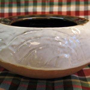 May include: A white ceramic bowl with a brown rim and a textured design. The bowl is sitting on a red and green plaid surface.